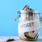 Glass jar filled with cash and labeled “Budget” against a blue background.