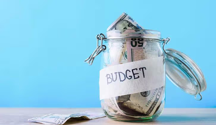 Glass jar filled with cash and labeled “Budget” against a blue background.