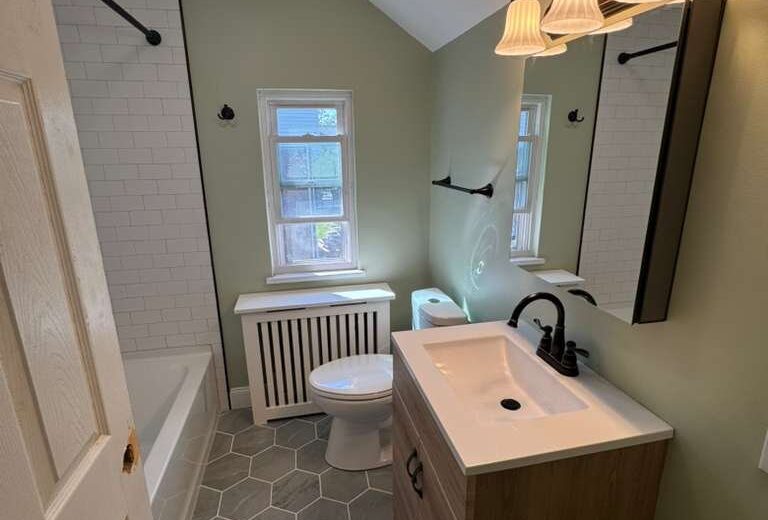 Bathroom vanity and toilet on gray hex tile with a window and white subway tub surround