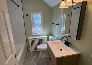 Bathroom vanity and toilet on gray hex tile with a window and white subway tub surround
